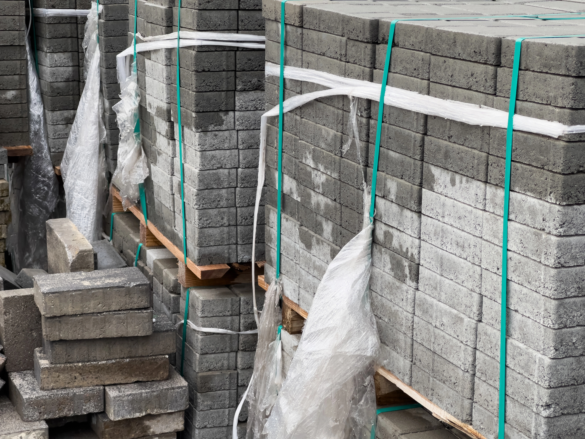 Stacks of gray concrete paving stones are secured with green and white plastic straps and partially covered with clear plastic sheeting.