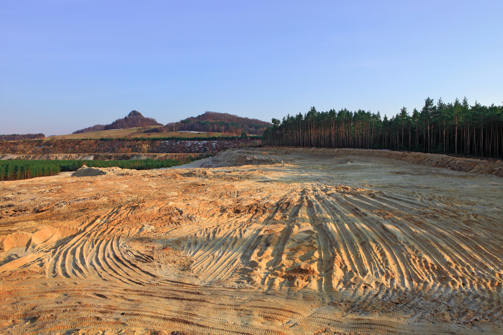 A large sandy area with visible tire tracks is cleared near a forest, with hills and a distant mountain under a clear sky in the background.