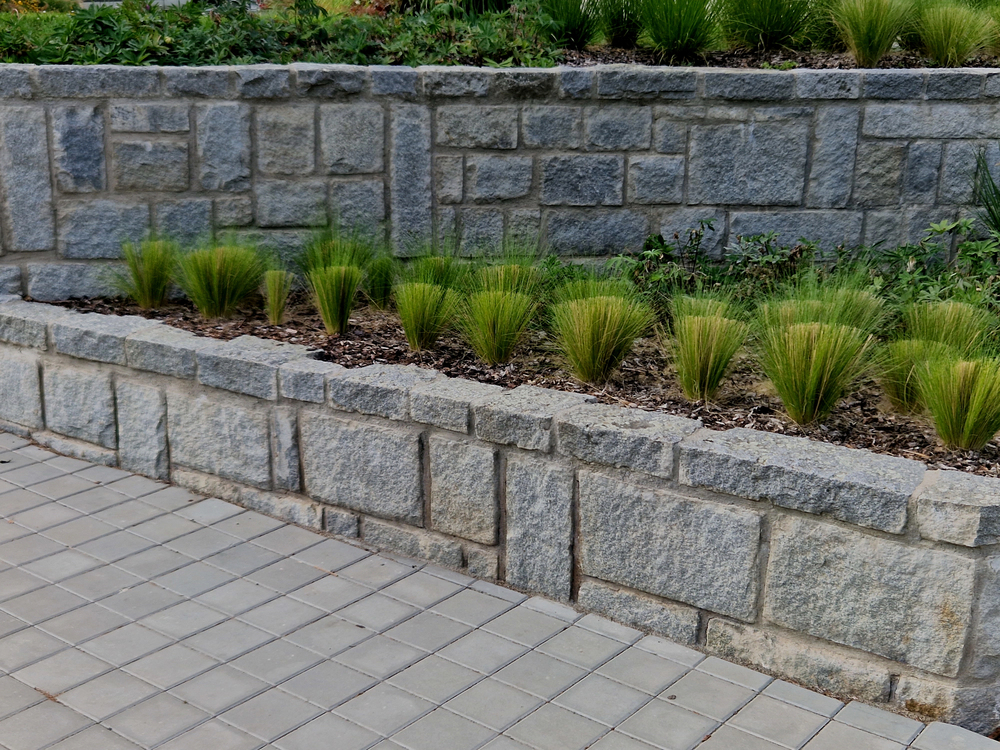 Stone retaining wall with rectangular tiles in the foreground and a row of ornamental grasses planted in soil along the base of the wall.
