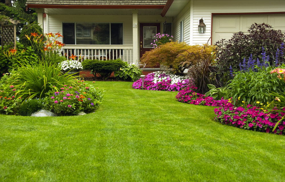 A well-maintained front yard with green grass, colorful flowerbeds, shrubs, and a porch in front of a white house.